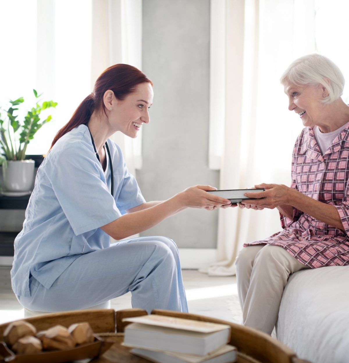 Nurse giving book. Caring nurse wearing uniform smiling while giving book to aged woman