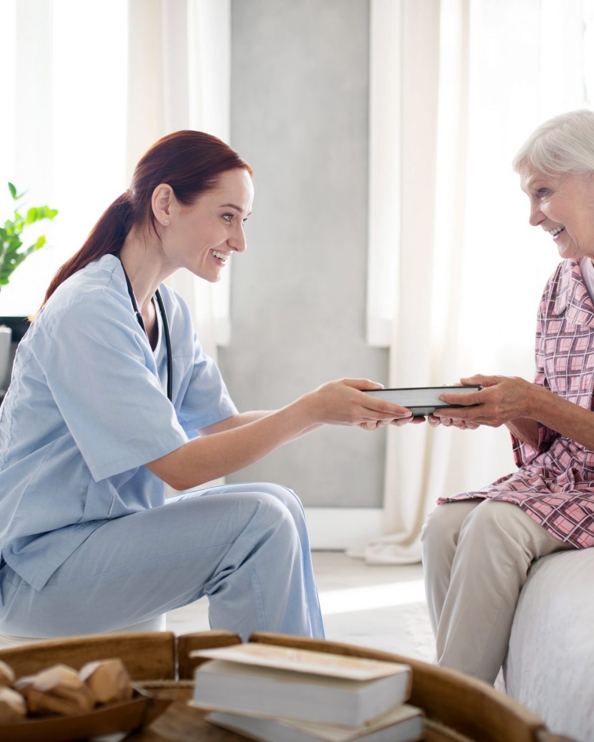Nurse giving book. Caring nurse wearing uniform smiling while giving book to aged woman